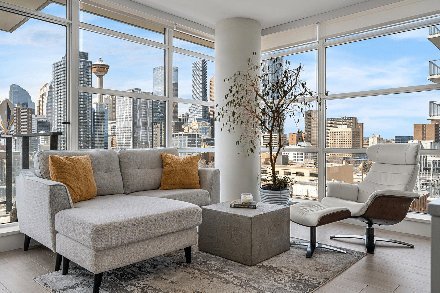 A modern living space featuring a gray sectional couch with orange cushions, a concrete coffee table, and a white recliner chair. Large windows provide a view of a city skyline.