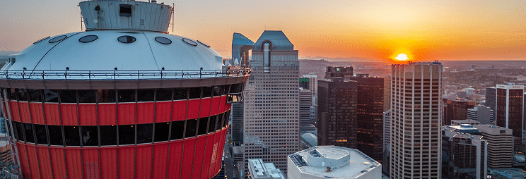 The image shows a city skyline at sunset, featuring a prominent tower with a circular, red-painted top and modern skyscrapers in the background.