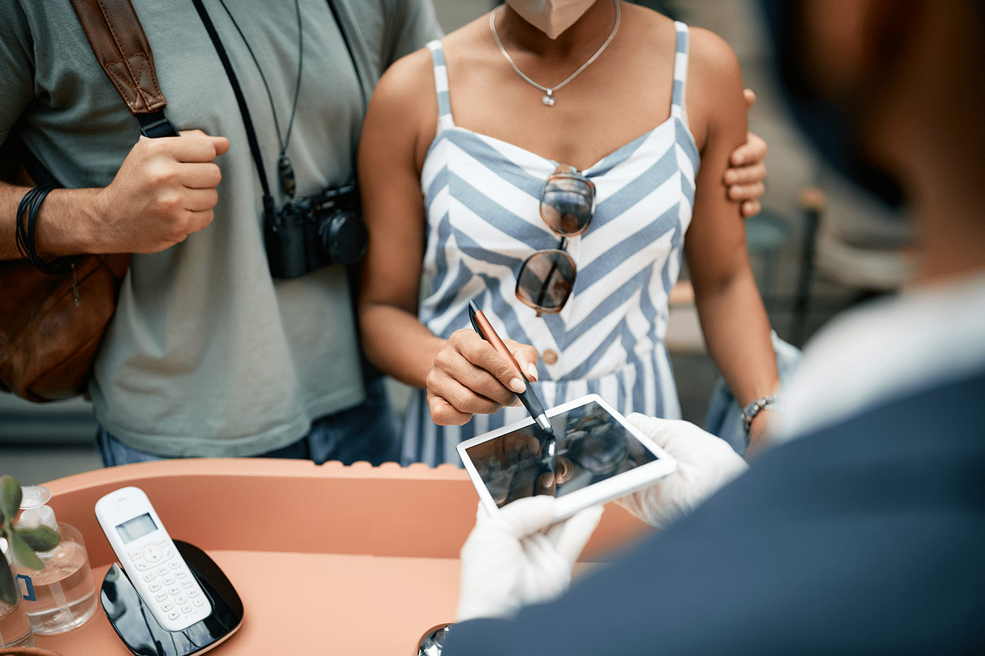 A woman in a striped dress is using a stylus on a tablet held by a person in gloves, while a man with a camera stands nearby.