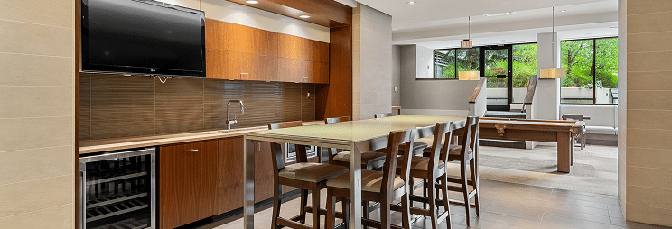 A modern kitchen and dining area with a large table, wooden cabinetry, a television, and a view of the outdoors through large windows.