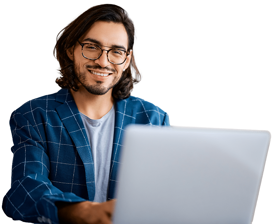 A cheerful man with long hair and glasses is sitting in front of a laptop, wearing a blue plaid blazer and a gray shirt.