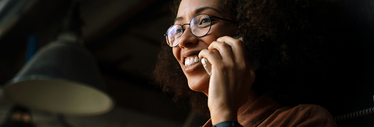 A smiling woman with curly hair and glasses is talking on the phone.