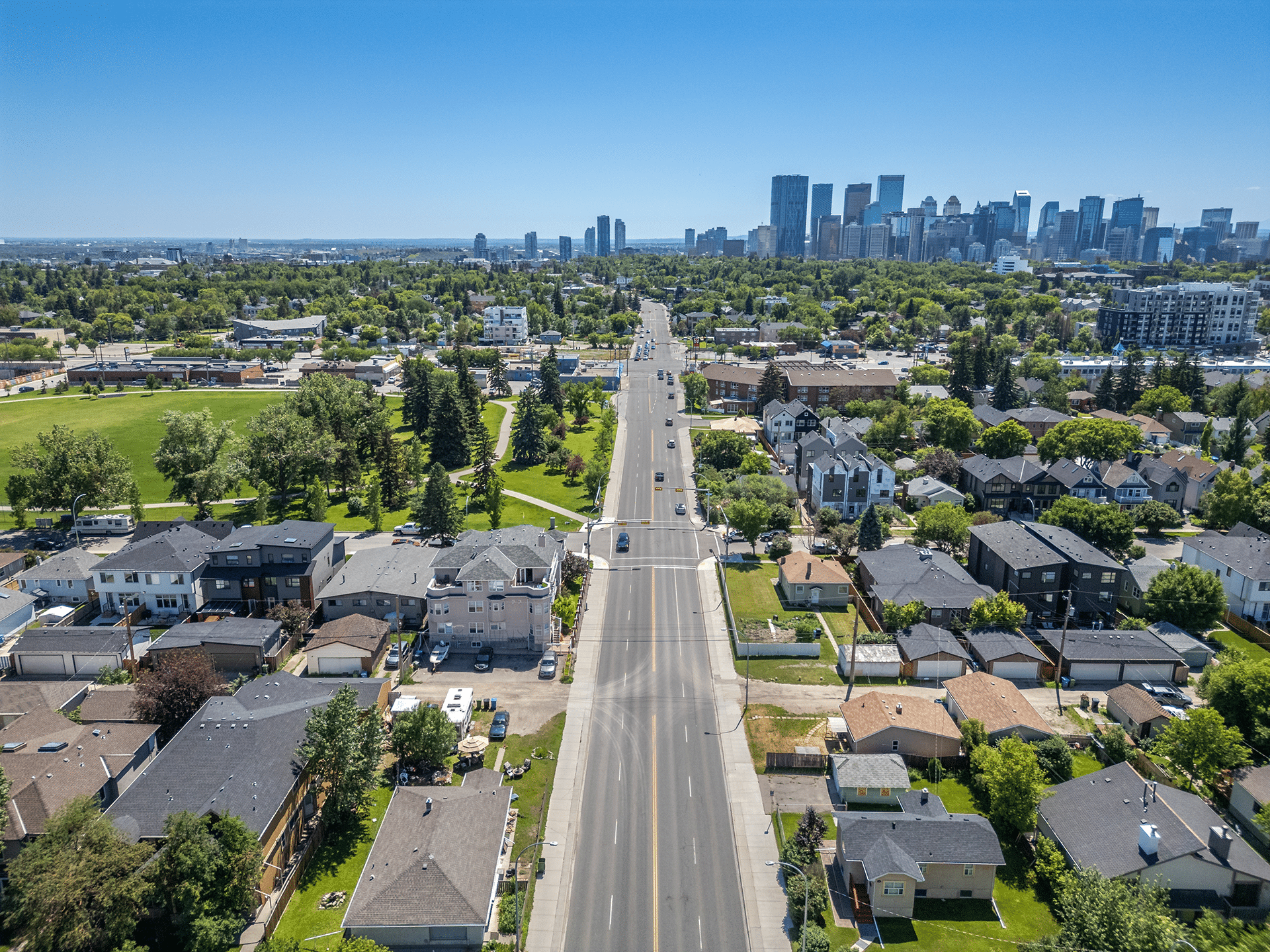 Aerial view of a residential neighborhood with houses, a green park, and a city skyline in the background under a clear blue sky.