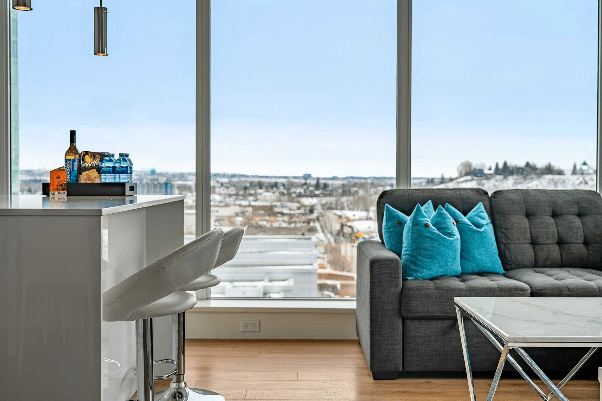 A modern interior featuring a gray sofa with turquoise pillows, a white bar counter, and tall stools. There are drinks and snacks on the counter, with large windows offering a view of a snowy city landscape.